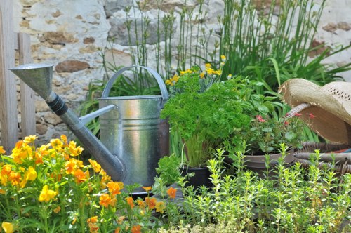 The image shows a lush, well-maintained garden corner with a variety of plants and gardening tools. In the foreground, there is a dense patch of vibrant yellow and orange flowers, likely marigolds, adding bright colour contrast to the scene. Behind these flowers, a weathered, metallic watering can sits on the ground, partially surrounded by tall green grasses or ornamental plants with slender leaves. To the right, a potted herb or leafy shrub with lush green foliage is visible, situated amidst other potted plants and garden elements. A beige sun hat rests on the surface near the pots, suggesting outdoor gardening activity. The background features a stone wall with a rustic appearance, and some additional greenery can be seen, including bushy plants and tall grasses. The scene appears to be set during daylight hours with natural lighting, evoking a peaceful and productive outdoor environment typical of a garden in Fulham, with careful arrangement showcasing typical gardening and outdoor maintenance elements.